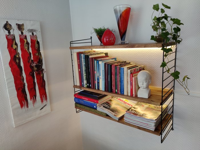 Stringhylla med hyllplan av ek, böcker och dekorativa föremål, mot vit tapetserad vägg.