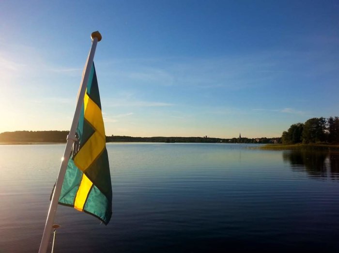 Svensk flagga i förgrunden med en lugn sjö och skog i bakgrunden under en klarblå himmel.