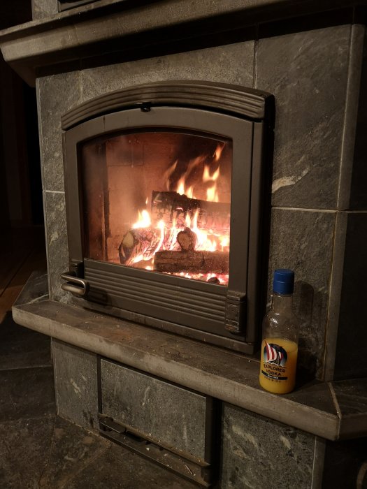 Tänd brasa i Tullikivi täljstenskamin med panorama-glasluckor, flankerad av stenplattor.