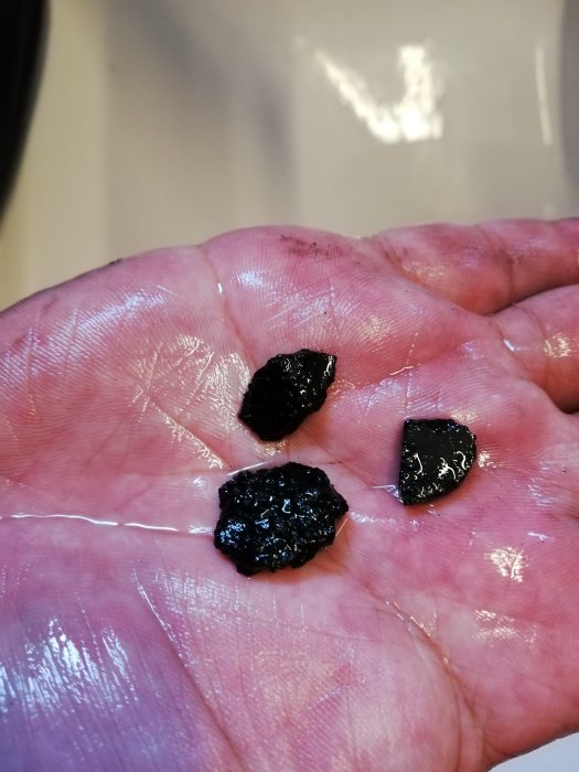 Handflata som visar tre bitar av svart, hårt material mot en suddig bakgrund, vilket antyder en byggrelaterad substans.