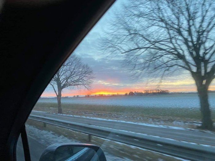 Soluppgång med glödande himmel, sedd från en bil mellan Lomma och Lund, med träd och snöigt fält.