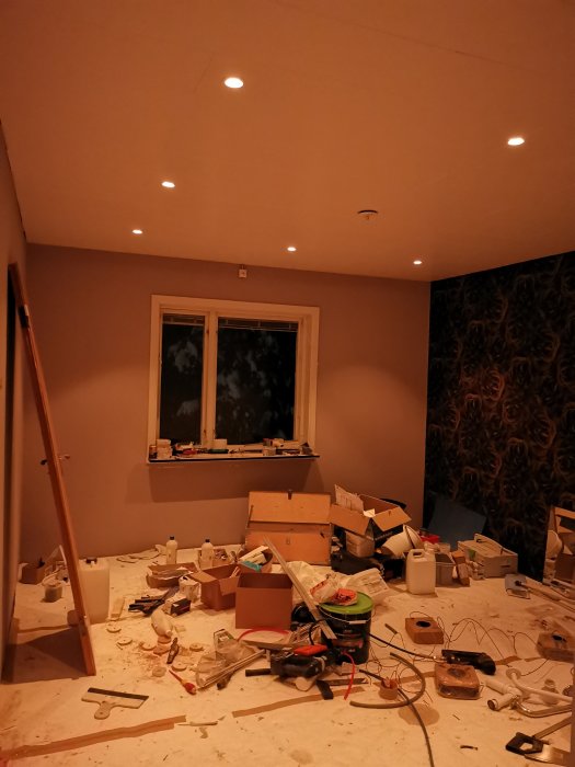 Rum under renovering med tända takspotlights, rörigt med byggmaterial och verktyg på golvet, fönster i bakgrunden.