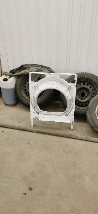 Demonterad torktumlare med öppen framdel i ett garage, omgiven av bildäck och behållare.