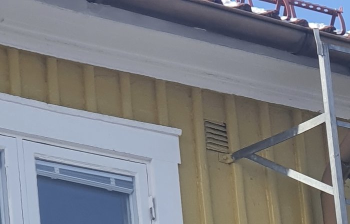 Ventilationsöppning nära takfoten på en gul vägg med en trappa och fönster.