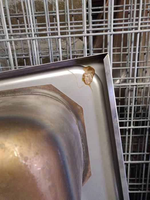 Närbild på en skada med brunaktig rest på kanten av ett silverfärgat köksredskap framför en metallgallerbakgrund.