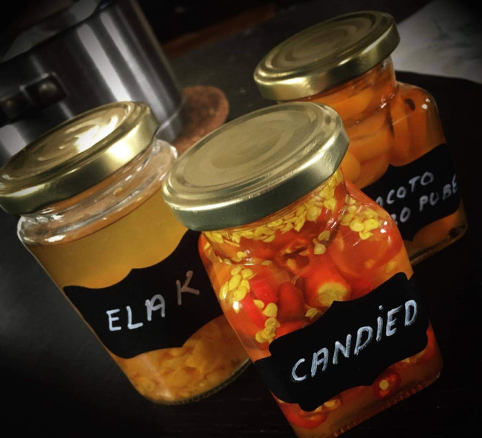 Tre glasburkar med etiketter, en märkt "ELAK" och en "CANDIED", fyllda med konservade varor på ett mörkt underlag.