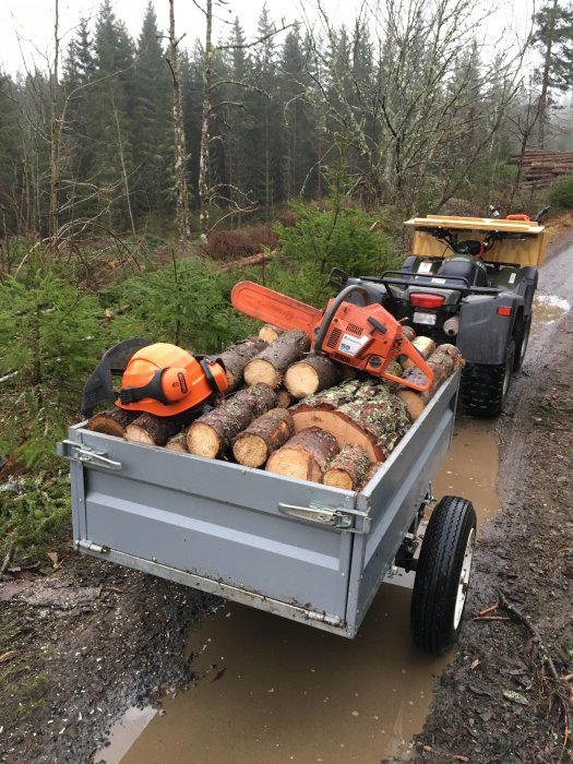 En ATV-kärra full med avkapade trädgrenar och en avställd motorsåg och hjälm på fuktig skogsväg.