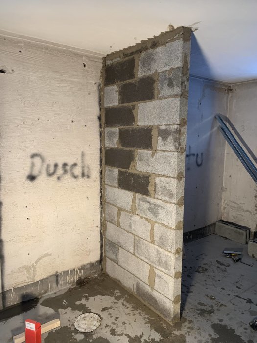 Renoveringsarbete med en oputsad tegelvägg, hantverksverktyg på betonggolv och texten "Dusch" sprayad på väggen.