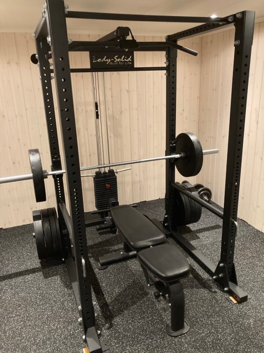 Nyinstallerat power rack för styrketräning med skivstång och viktskivor, komplett med latpull-tillbehör och bänk, i ett hemmagym.