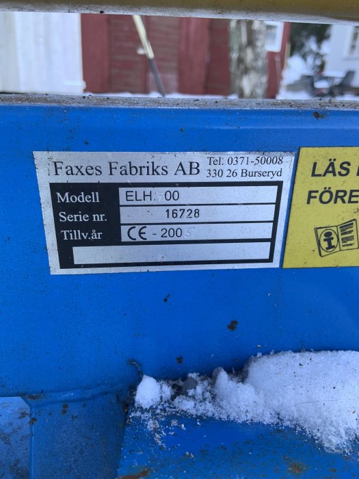 Informationsdekal på en vedklyv från Faxes Fabriks med modellnummer och tillverkningsår, täckt delvis av snö.