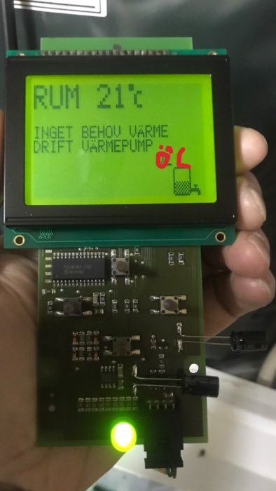 Hand håller en elektronisk kretskort med en fungerande display som visar "RUM 21°C INGET BEHOV VÄRME DRIFT VÄRMEPUMP".