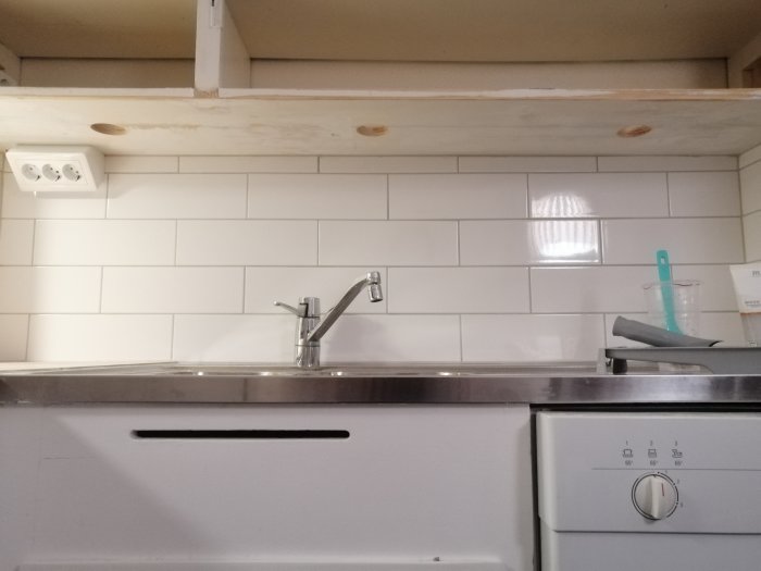 Kök med vit kakelvägg och förberedda hål för installation av downlights över diskbänk och spis.