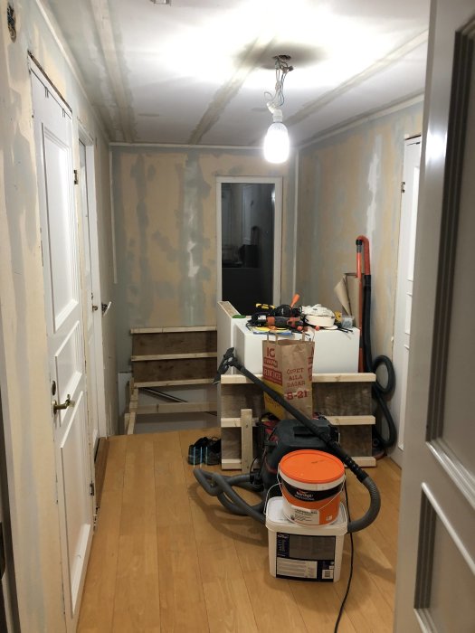 Korridor under renovering med spacklade väggar och tak samt verktyg och byggmaterial på golvet.