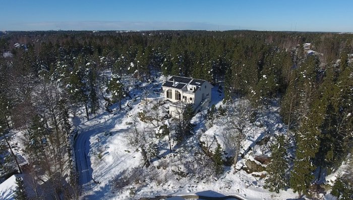 Hus med solpaneler på taket omgivet av snötäckt skog och klarblå himmel, sett från luften.