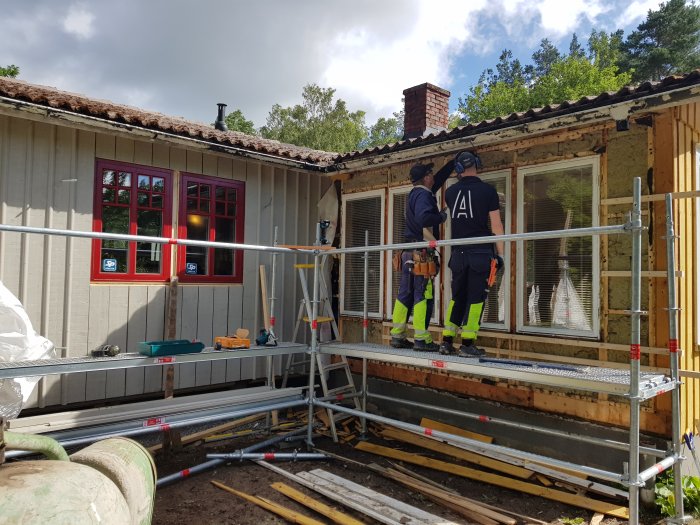 Två personer installerar ett fönster i ett hus under renovering, med byggmaterial och ställningar synliga.