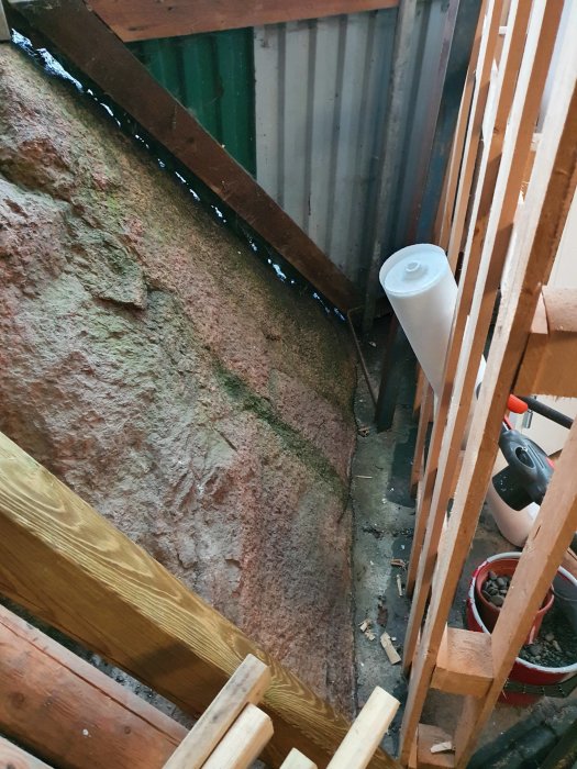 En hörna av ett trädgårdsförråd med korrugerad plåt och en synlig bergvägg, indikerande fuktproblem och renoveringsbehov.