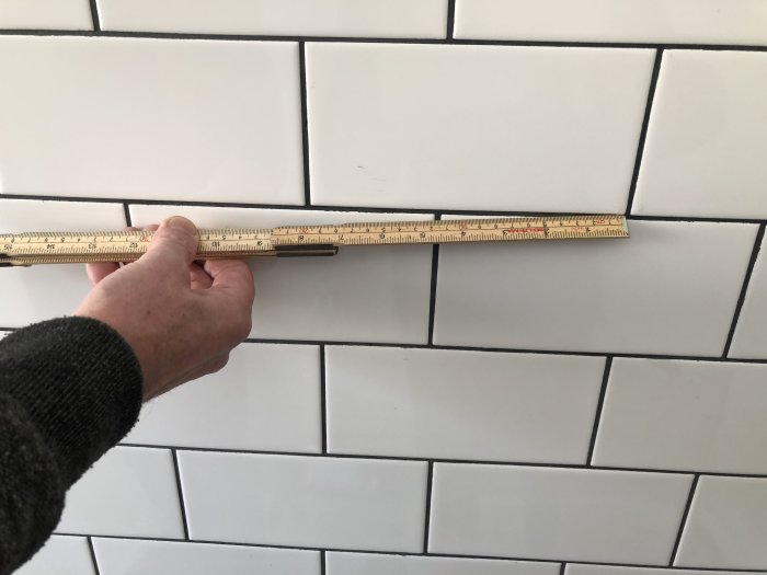 Hand som håller en tumstock mot en kaklad vägg för att mäta plattornas placering.