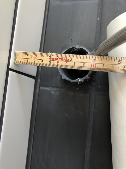 Måttband mäter avstånd på en kakelplatta nära en golvbrunn med synliga rör och kablar.