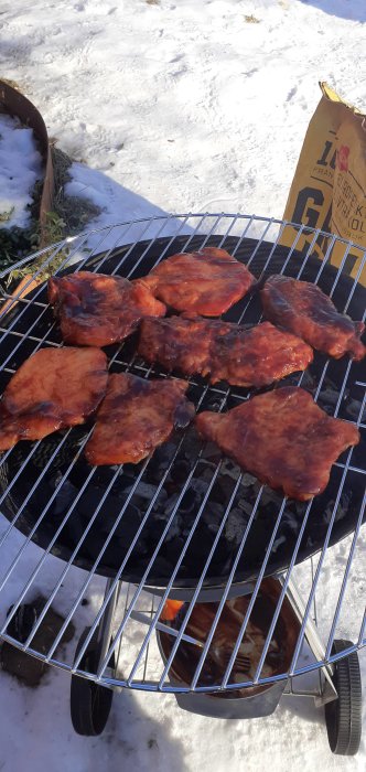 Grillade revbensspjäll på en kolgrill utomhus med snötäckt mark i bakgrunden.