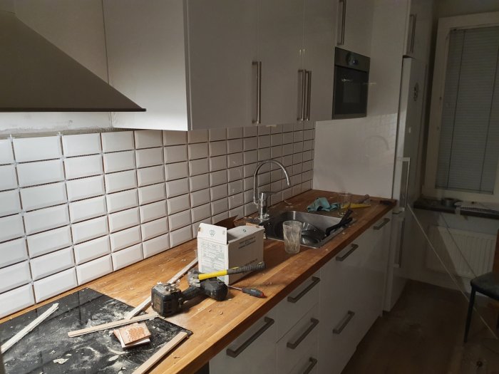 Kök under renovering med verktyg och byggmaterial på träbänkskiva och vita kakelväggar.