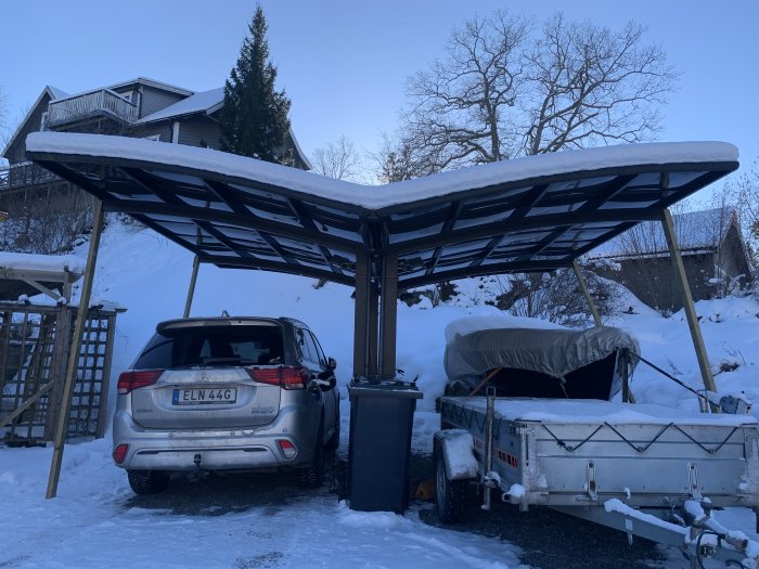 Carport med snötäckt tak som böjer sig under vikt, bil och släpvagn parkerade under.