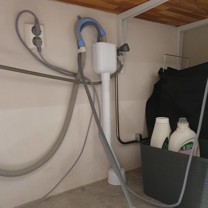 Vattenanslutningar och avloppsslang bakom en tvättmaskin med tvättmedel i korgen.