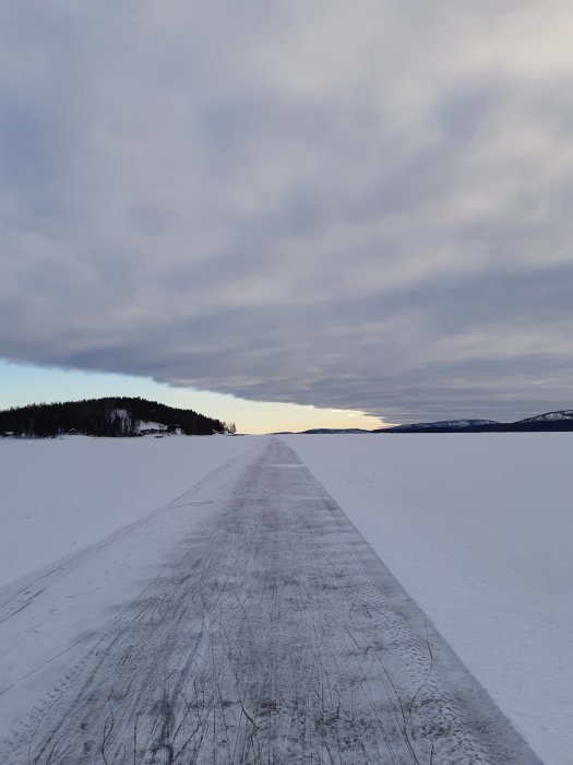 Snötäckt isväg på en frusen sjö med molniga himmel och antydan till solnedgång i fjärran.