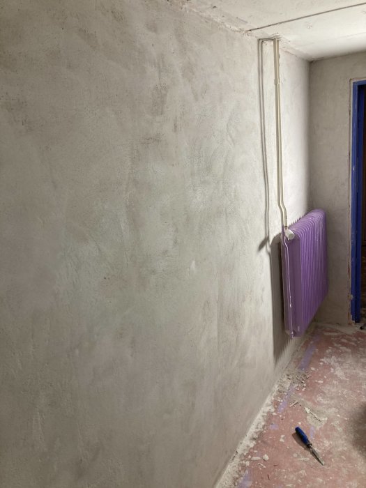 Nyputsade väggar i en källare med en lila radiator, blå dörr och ett verktyg på golvet.