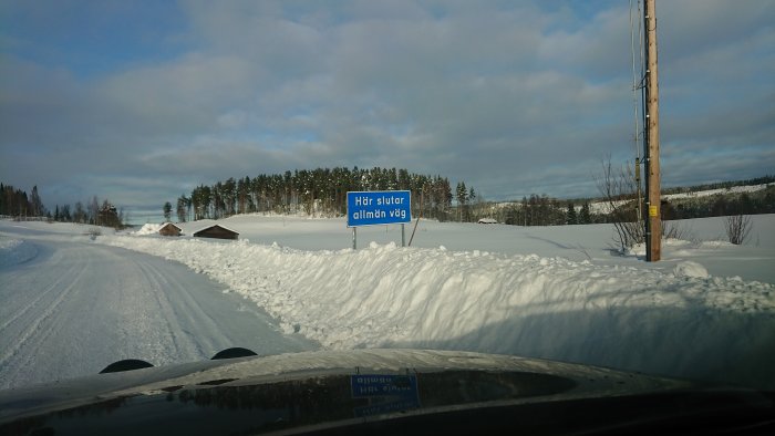 Vinterlandskap med skylt som säger "Här slutar allmän väg" vid en snötäckt väg med bilens huva i förgrunden.