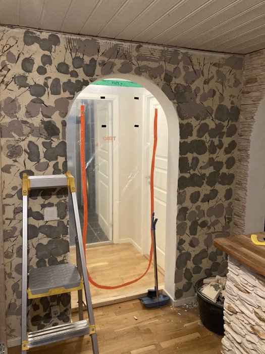 Putsad vägg med inristade grenavtryck i ett rum med stege och skyddsplast vid dörröppning.