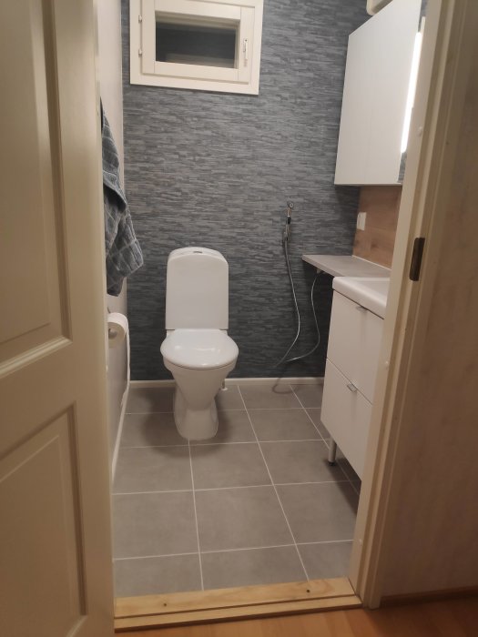 Renoverat toalettrum med grå klinker, grått kakel på väggen, vit toalett och handfat, och handdukshängare.