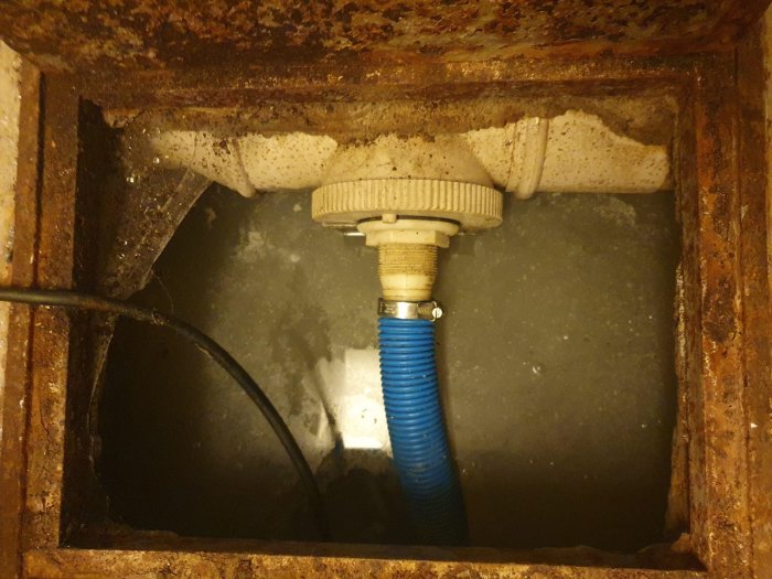 Vy inifrån en rostig pumpgrop med en blå slang ansluten till pump, tydliga avlagringar och vatten i källarmiljö.