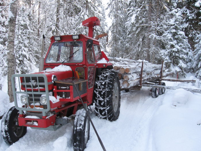 Röd skogsmaskin med timmersläp lastat med stockar på en snötäckt skogsväg.