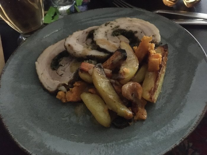 Långstekt porchetta med en fyllning av gröna örter och serverad med ugnsstekta grönsaker som potatis och morötter.