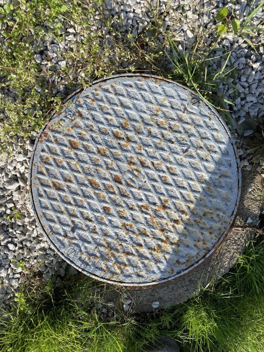 Rostig brunnslock av metall omgiven av grönt gräs och grå småsten.