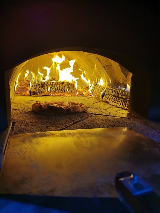 Pizza bakas i vedeldad ugn med flammor längs bakväggen och vedträn till vänster.