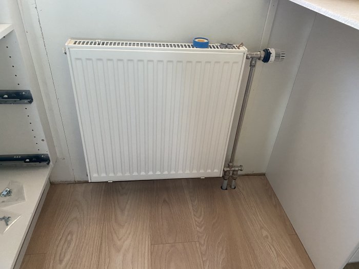 Vit radiator framför en vägg med synliga rör och inbyggnadsskenor för skåp ovanför.