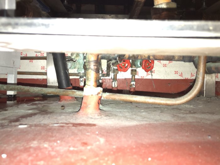 Vy under ett golv som visar rör och ventiler avsedda för husets värmesystem, inklusive en röd skruvventil.