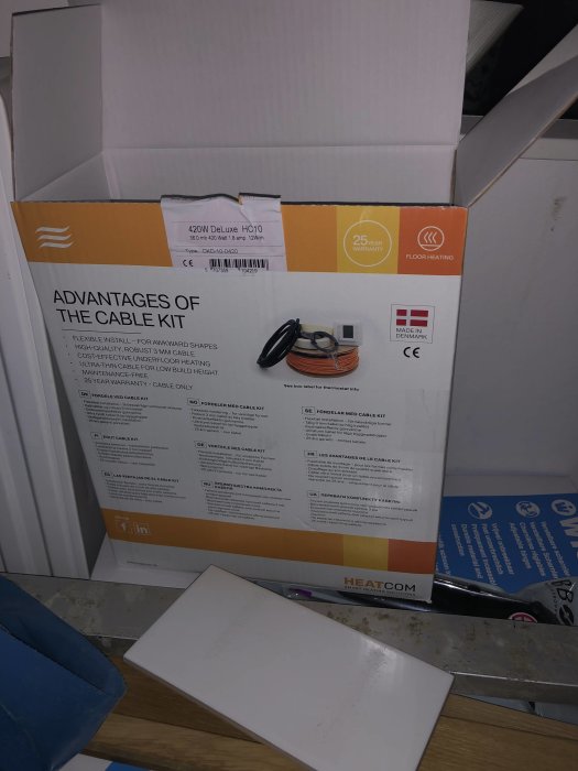 Öppnad låda med golvvärme kit från HEATCOM, inklusive en rulle med värmekabel och en installationsmanual, mot en bakgrund av byggmaterial.