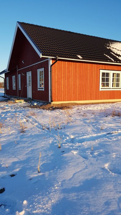 Röd trävilla med snötäckt mark i vintersol, plats för framtida altan markerad i snön.