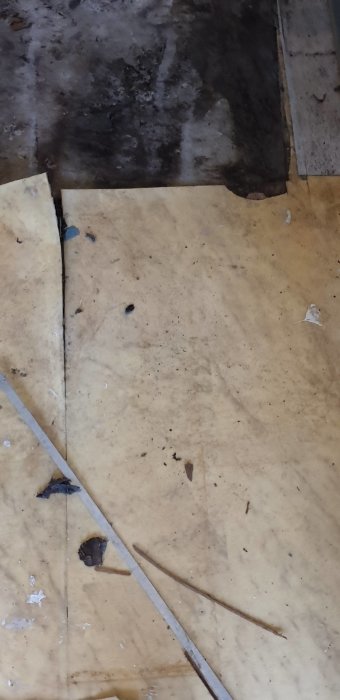 Gammalt och skadat golv med fläckar och vattenskador, delvis täckt av gul plastmatta.