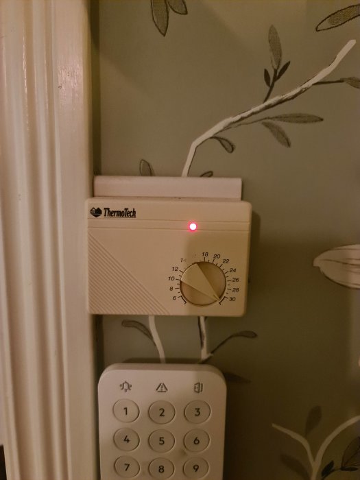 En Thermotech termostat med vred och lysande röd indikator monterad på en vägg med blommigt tapetmönster.