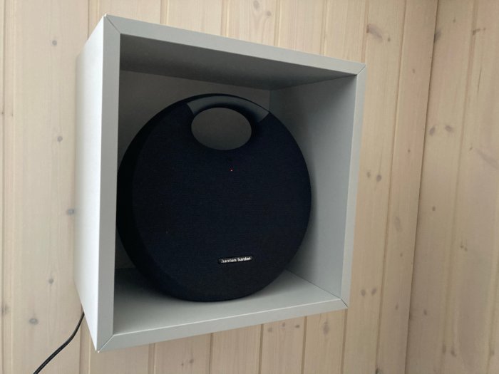 Harman Kardon Onyx Studio 6 högtalare på en vit hylla mot en trävägg.