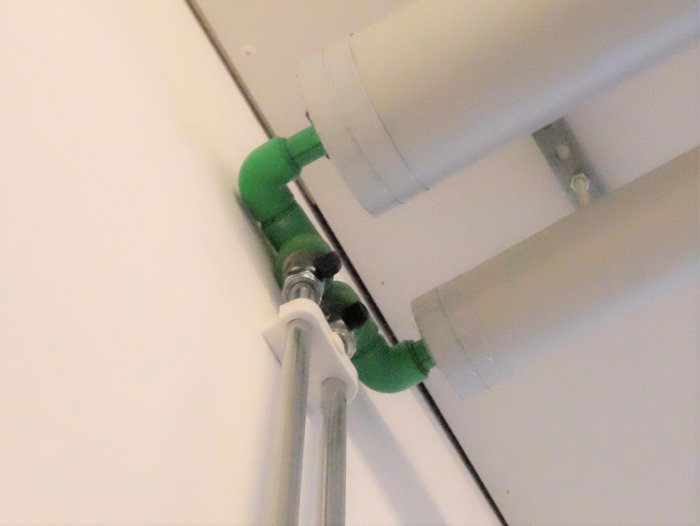 Koppling av grönt plaströr till kopparrör med rördelar och fästen mot en vit vägg.