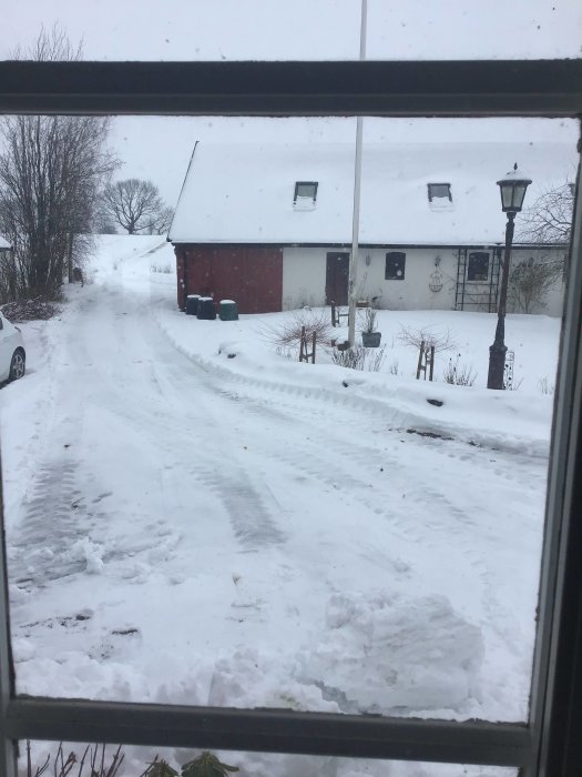 Vy genom fönster på nyplogad innergård täckt med snö, hus och bil i bakgrunden.