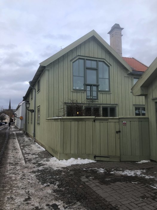 Tvåvåningshus med ljusgrön färg och vitt tak under grå himmel, omgiven av snötäckt mark och kullersten.