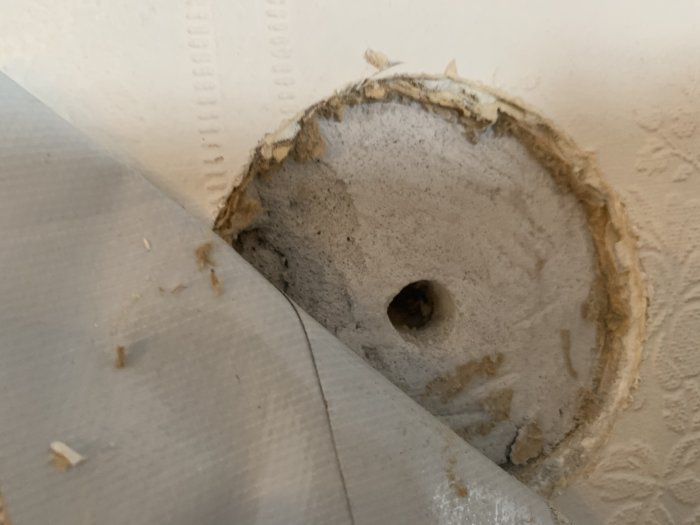 Inspektionshål visar en grå skiva fastsatt direkt på en murstock med tapetresidue runt kanterna, misstänkt för asbest.