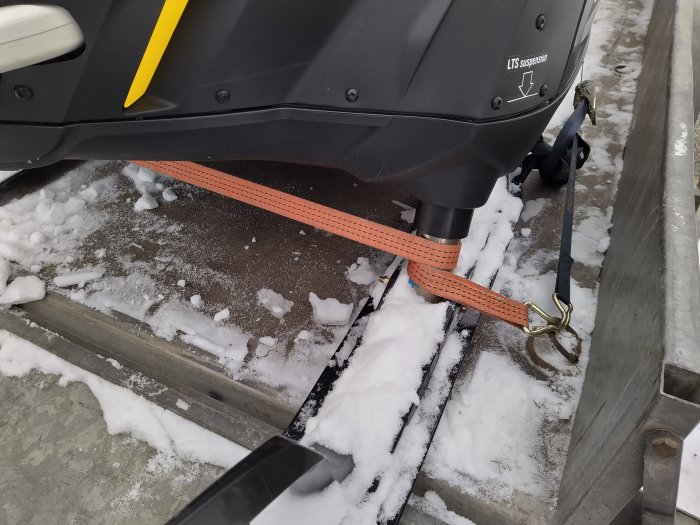 Spännband säkrar en snöskoter vid teleskop-benen, fastsatt vid ett släp, omgiven av snö och en grå ramp.