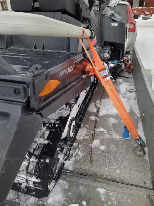 Skoter säkrad på släp med orange spännband runt framdel och snö på marken.