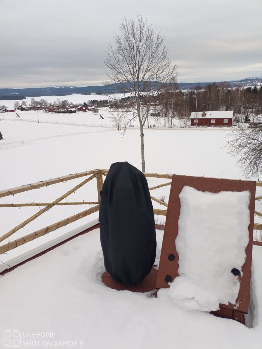 Teleskop under skyddande överdrag på en snöig terrass med utsikt över ett vinterlandskap.